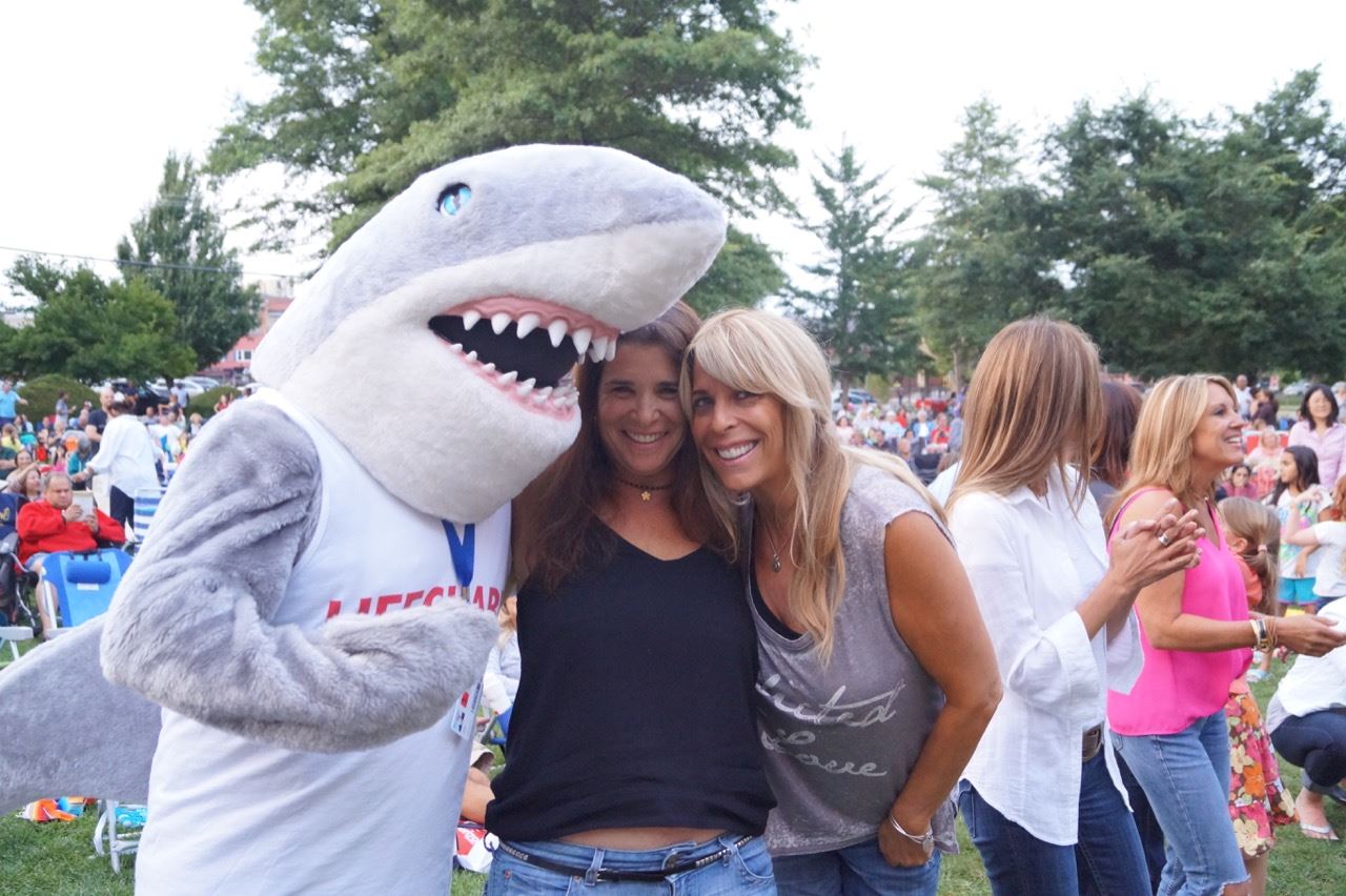 The shark mascot poses with 2 women in the crowd.